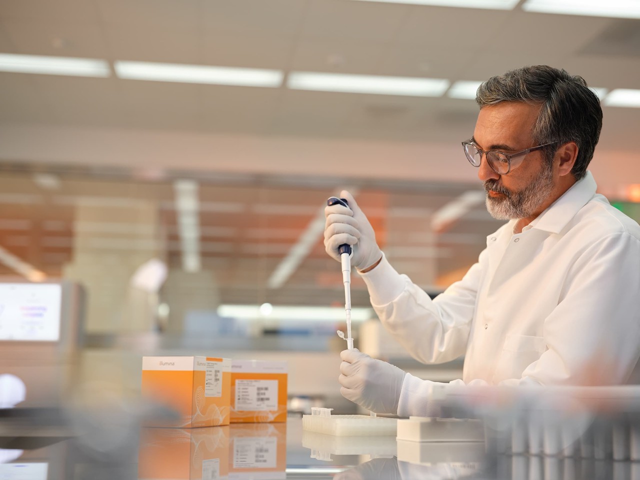 Male scientist is using a single pipette into a tube, blurry images of a NovaSeq X and TruSight Oncology ctDNA v2 Enrichment library prep boxes in the background. 