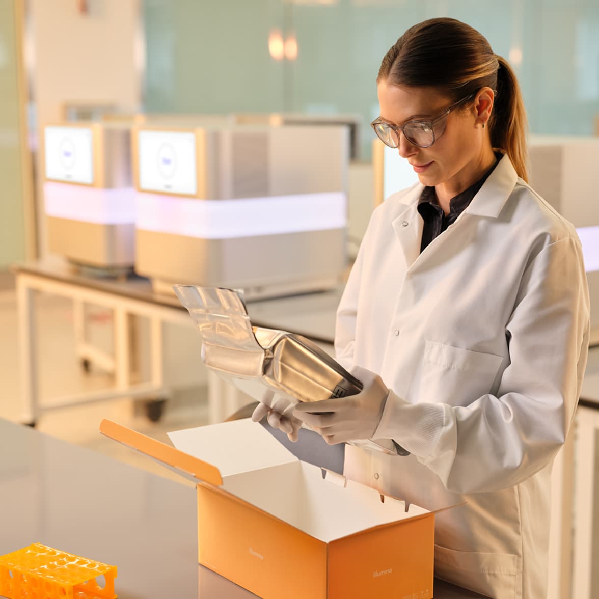Front view of female scientist holding and inspecting foil wrapped NextSeq 1000/2000 reagent cartridge, pulled out from the box; multiple NextSeq 1000/2000 instruments blurry in the background.