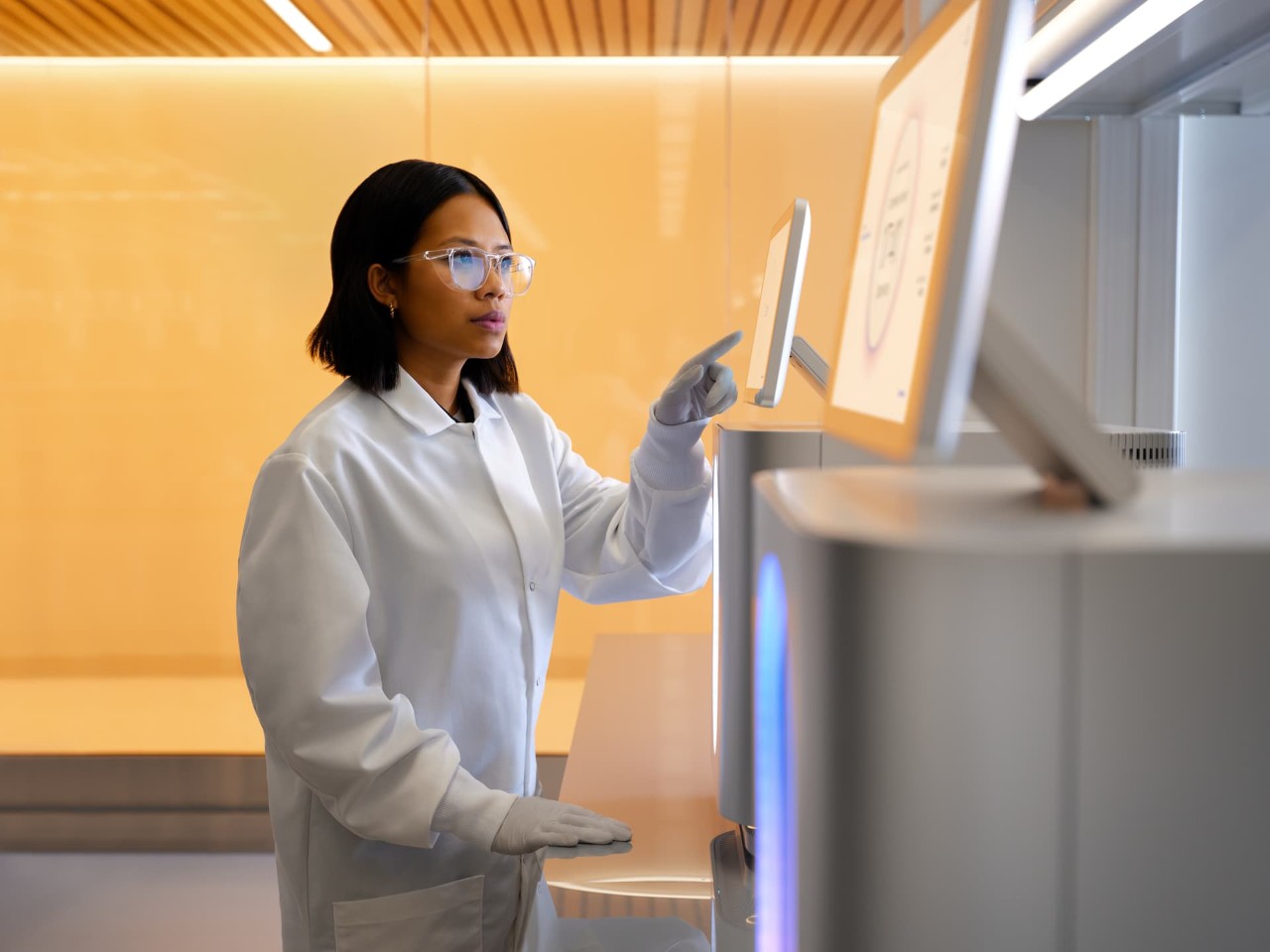 Female scientist using finger to make a selection from the start touch screen monitor while looking at the finish screen on another instrument. Angled side view of person and instruments on lab bench.