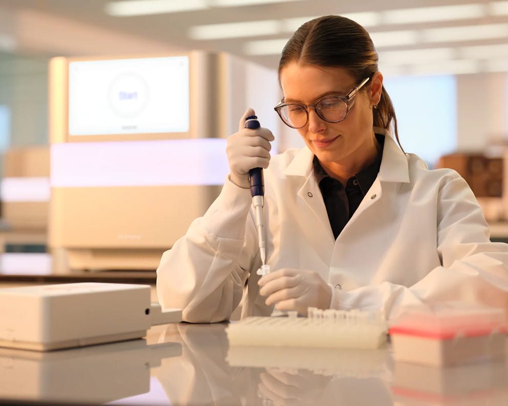 Front view of a female scientist pipetting clear liquid into tube; reagent cartridge, box, and plates in the foreground; NextSeq 1000/2000 instruments in the background.